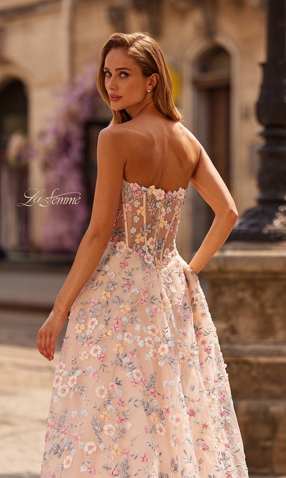 A woman outdoors, looking over her shoulder in the La Femme 33409: a long prom dress featuring a strapless sweetheart neckline, floral embroidery, and sheer back—an elegant designer gown for special occasions.
